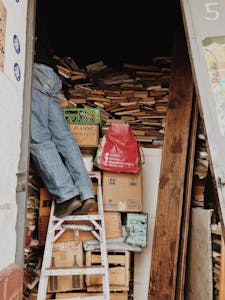 A person climbing a ladder in a cluttered storage area filled with books and boxes.